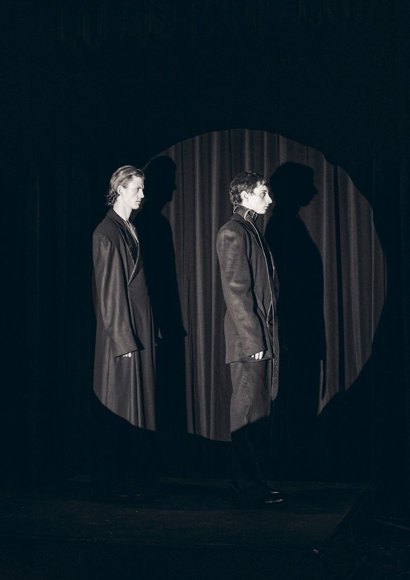 Two adult figures stand in front of a neutral backdrop. The figures are lit with hard lighting casting stark shadows behind them.