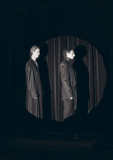 Two adult figures stand in front of a neutral backdrop. The figures are lit with hard lighting casting stark shadows behind them.