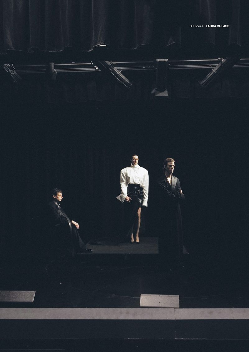 Three fashion models in dark clothing pose on a dark stage with black backdrop and metal lighting equipment at a fashion show.