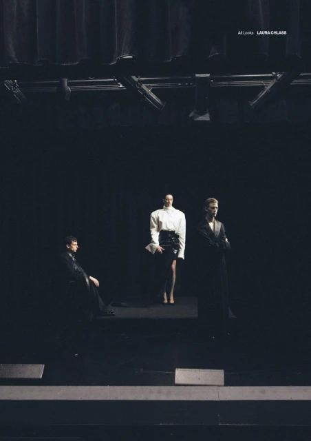 Three fashion models in dark clothing pose on a dark stage with black backdrop and metal lighting equipment at a fashion show.