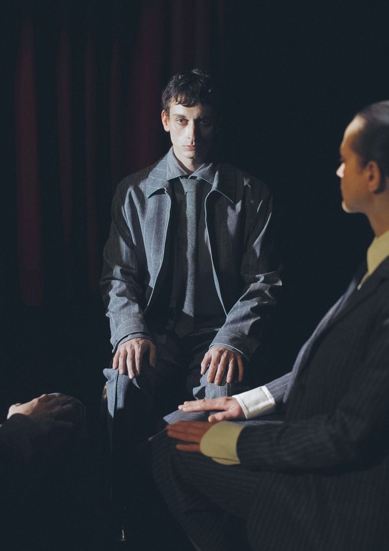 Two men in suits are positioned in a darkly lit indoor setting, possibly a stage, with focus on the seated man in gray attire.