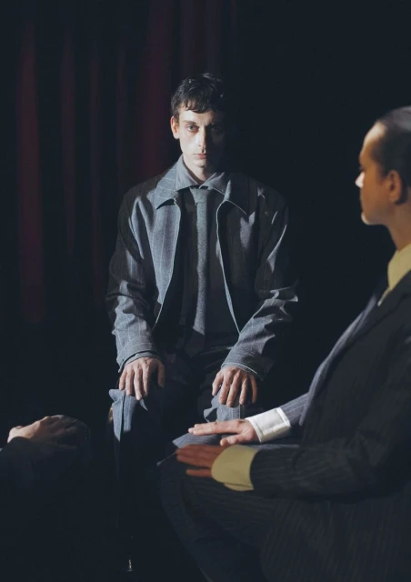 Two men in suits are positioned in a darkly lit indoor setting, possibly a stage, with focus on the seated man in gray attire.