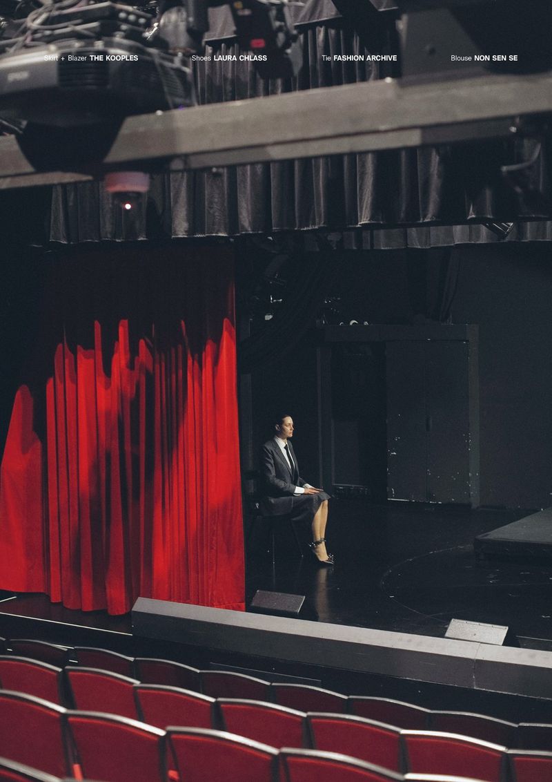 Elegant woman in a suit sits on a stage with red curtains and rows of empty red seats, under bright stage lighting.