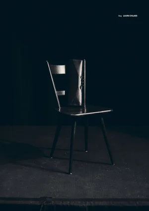 A dark wooden chair standing on a dark surface against a deep black studio background with a clutch bag resting on the seat