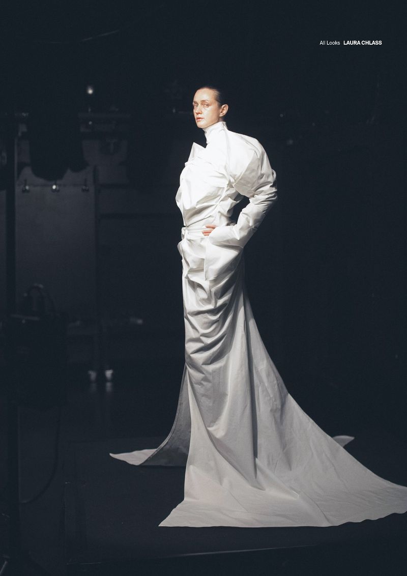 Model in a voluminous white structured gown with a high collar and long train standing against a dark background.