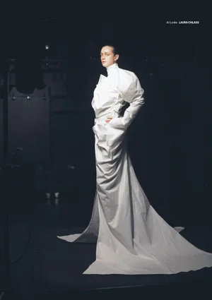 Model in a voluminous white structured gown with a high collar and long train standing against a dark background.