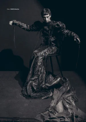 A dark-haired man seated on a chair in a dark studio wearing a textured black leather outfit with dramatic flowing fringes and a long draped hem.