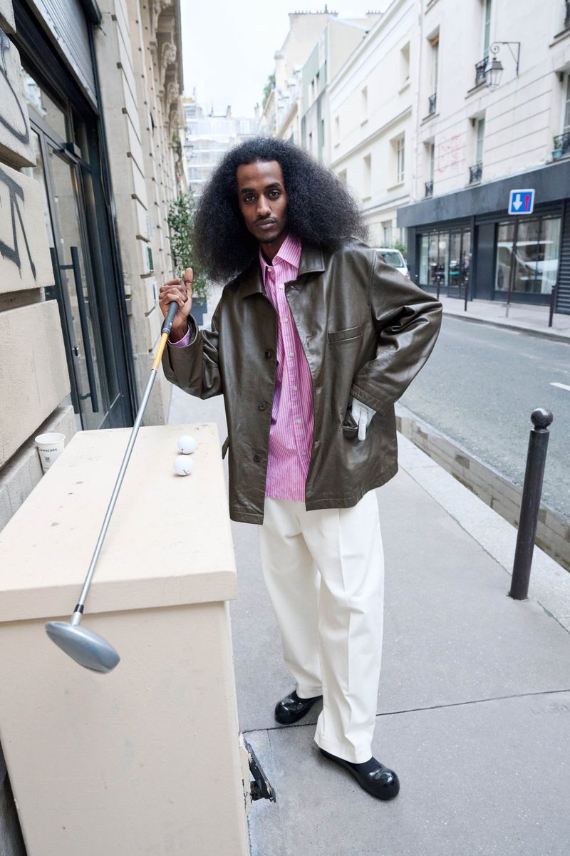 Stylish man in street with golf club, wearing a pink shirt and olive jacket. Street scene in soft light, architectural backdrop.