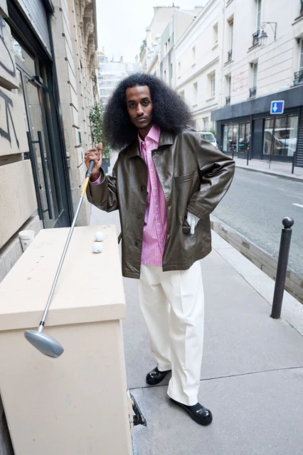 Stylish man in street with golf club, wearing a pink shirt and olive jacket. Street scene in soft light, architectural backdrop.
