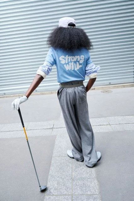An adult with a golf club, wearing a blue shirt with the text 'Strong Will' on it. The person is wearing gray pants and a white cap.