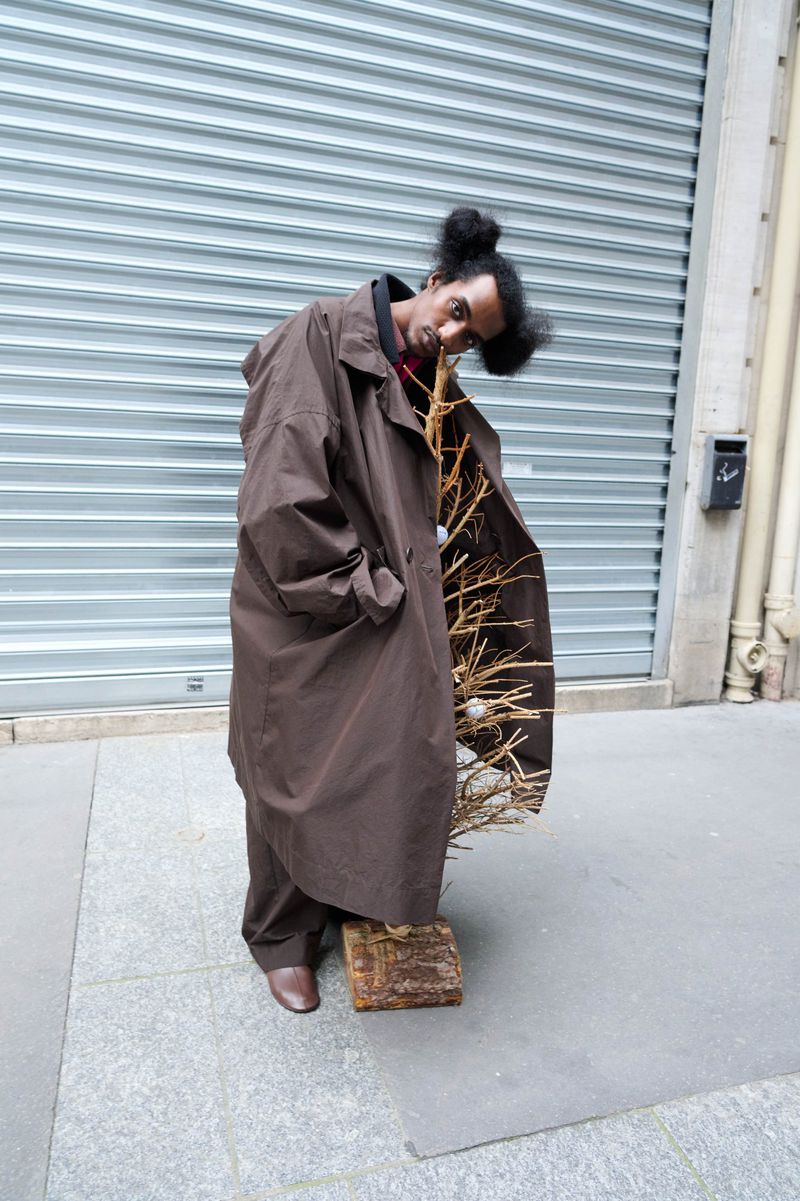 A person in a brown coat stands on a piece of wood holding bare branches, with a metal roll-up door in the urban background.