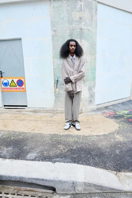 Young adult man in a tan suit stands on concrete with a sand patch in front of a weathered building with a closed door and hazard sign.