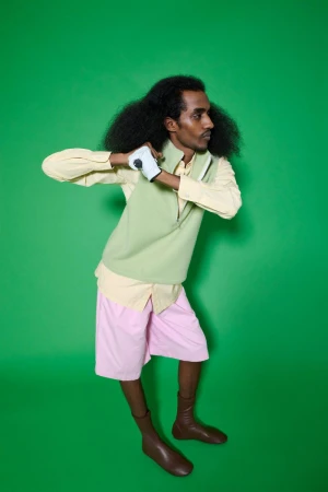 Man with long curly hair in golf attire poses in front of a green screen, wearing pink shorts, a light green vest, and brown boots.