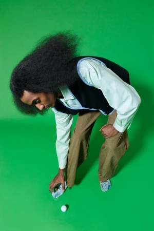 High-angle shot of a Black male in vest and slacks bending over to touch a golf ball on a bright green seamless backdrop in studio lighting.