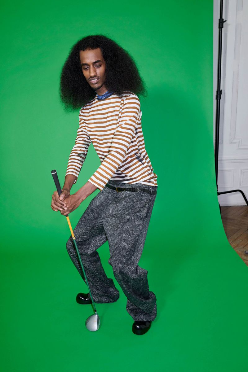A man with dark hair and striped shirt poised to swing a golf club against a bright green backdrop in a studio setting.