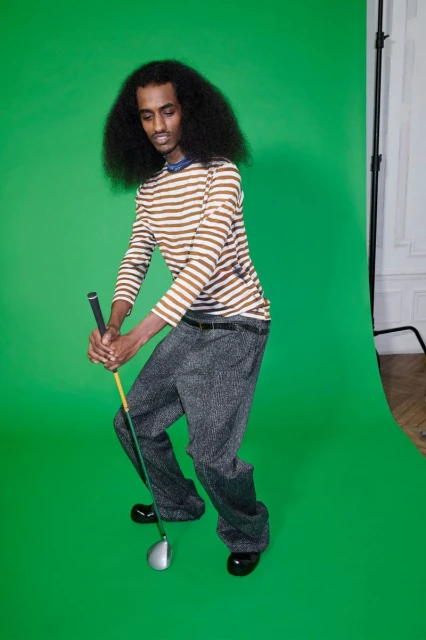 A man with dark hair and striped shirt poised to swing a golf club against a bright green backdrop in a studio setting.