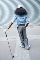 Model seen from behind holding a golf club while wearing a light blue printed long-sleeve shirt and grey trousers in front of a metal shutter