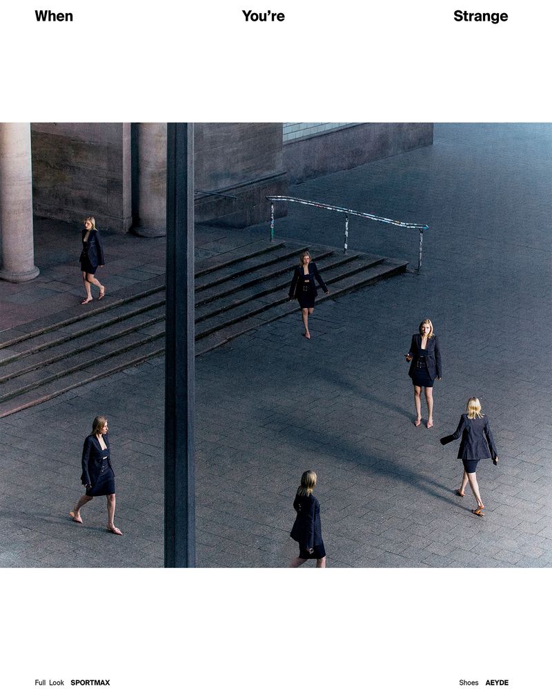 Fashion editorial shot of six women in black suit dresses walking across a large, gray concrete plaza and building exterior on a sunny day.
