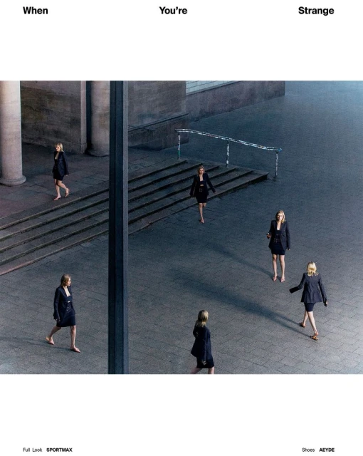 Fashion editorial shot of six women in black suit dresses walking across a large, gray concrete plaza and building exterior on a sunny day.
