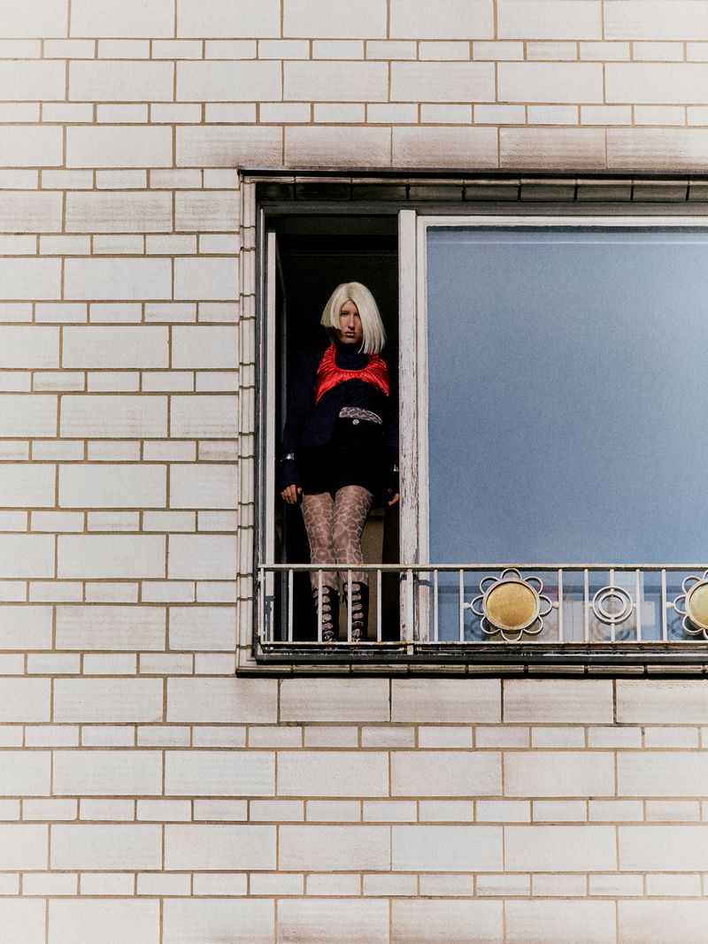 Young adult woman with blonde hair stands at a window wearing a red vest, black sweater, patterned tights, and laced boots.
