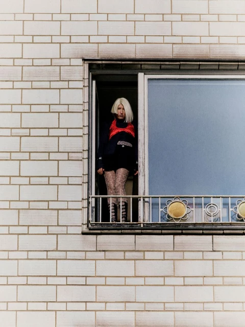 Young adult woman with blonde hair stands at a window wearing a red vest, black sweater, patterned tights, and laced boots.