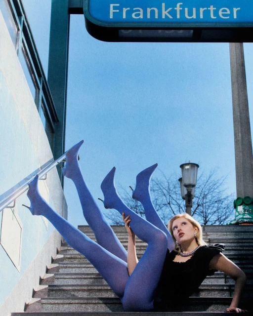 Woman in black dress and blue stockings poses on gray stairs under a sign reading "Frankfurter" against a blue sky background. 