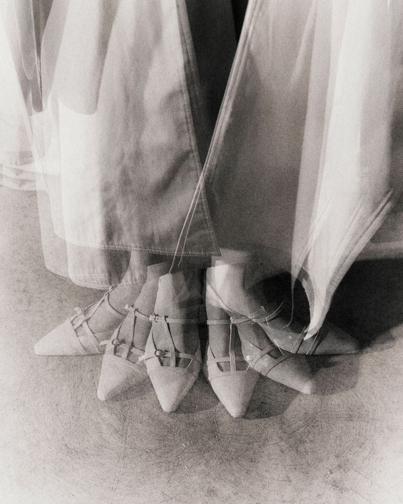 Low angle, monochrome shot of a person in a white dress with multiple exposures of white pointed shoes on a light grey surface.