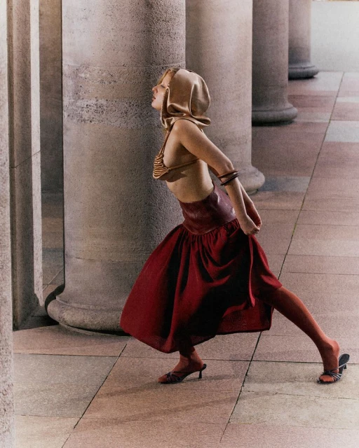 Woman in stylish hooded top, bra top, red skirt, and stockings poses between concrete columns in an outdoor fashion setting.