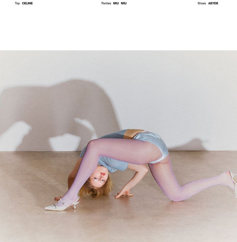 A woman in a blue top and lilac tights arches her back, hands on the floor, showcasing fashion clothing and accessories in a light setting.