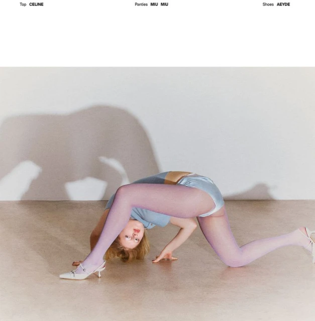 A woman in a blue top and lilac tights arches her back, hands on the floor, showcasing fashion clothing and accessories in a light setting.