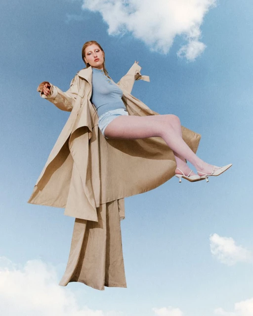 A model floats on a sky-blue background wearing a trench coat, light blue top, denim shorts and white heels in a studio fashion shot.
