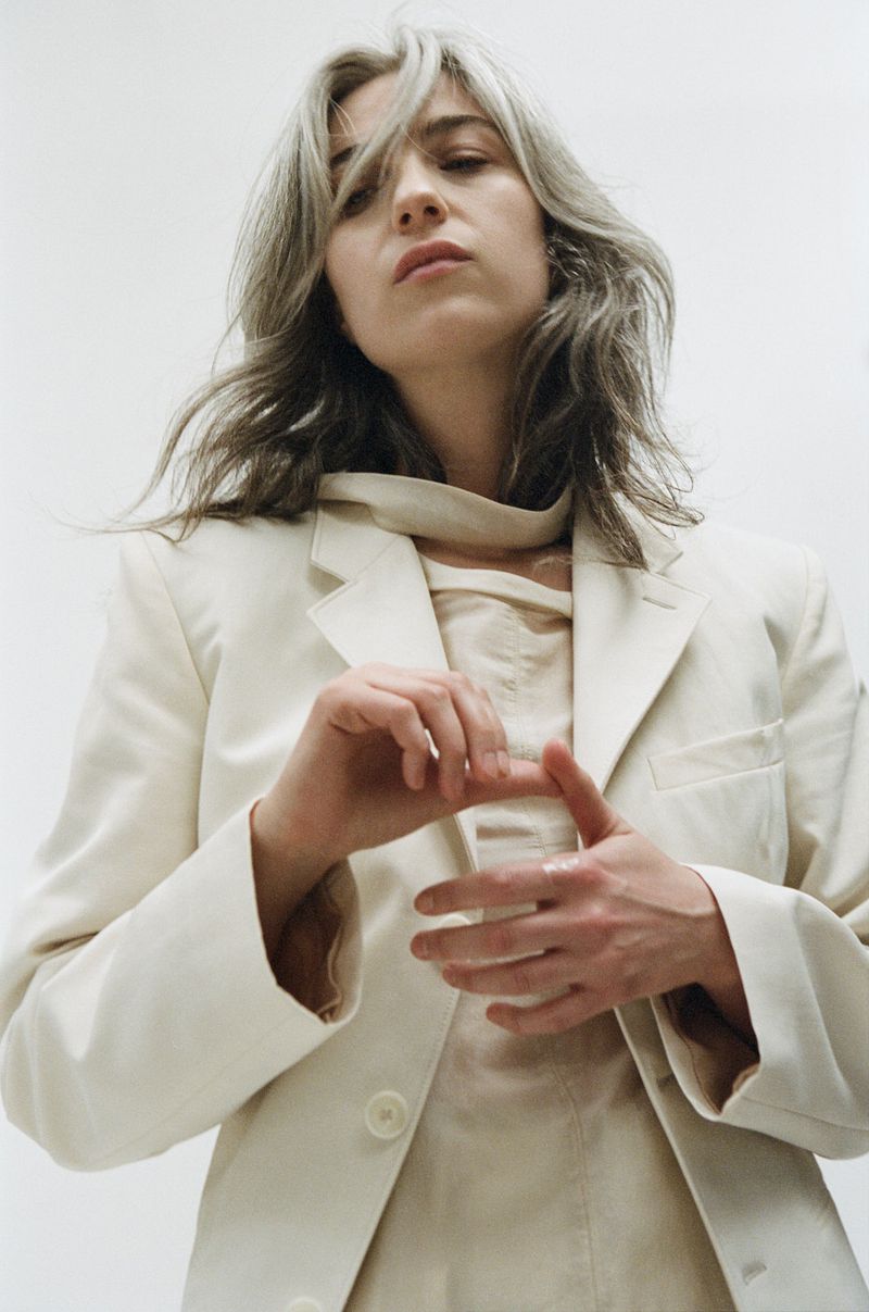 Portrait of an adult female with grey hair wearing a light-colored blazer and shirt, shot at eye-level in a bright, indoor studio setting.