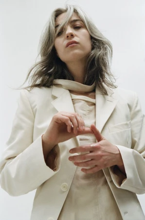 Portrait of an adult female with grey hair wearing a light-colored blazer and shirt, shot at eye-level in a bright, indoor studio setting.
