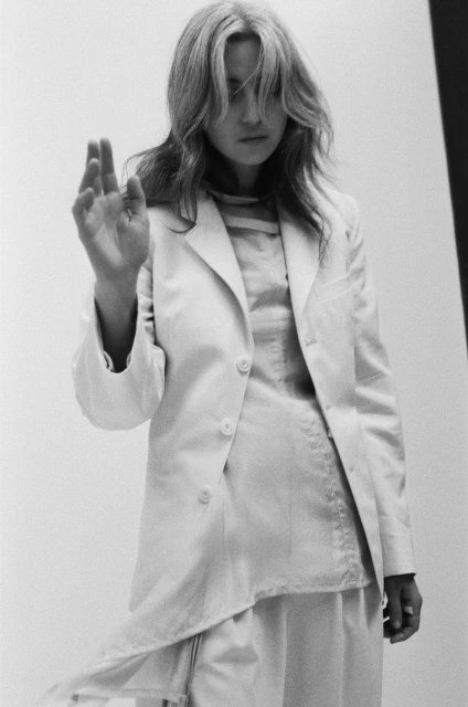 Woman in a white blazer and skirt stands in front of a white wall with one hand raised in a gesture, black and white photography.
