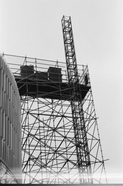 Black-and-white image featuring a tall, metal scaffold structure with containers on top, seen against a bright sky and part of a building.
