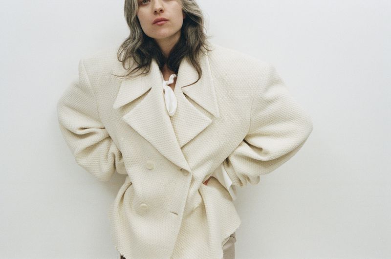 Woman in a textured cream-colored double-breasted overcoat stands against a white wall in a fashion shot with a neutral expression.