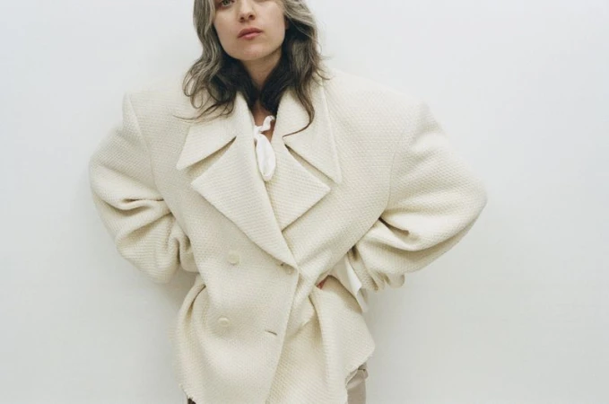 Woman in a textured cream-colored double-breasted overcoat stands against a white wall in a fashion shot with a neutral expression.