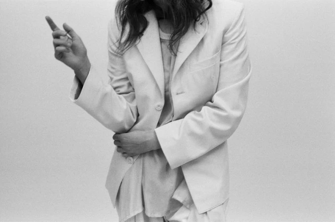 Monochrome portrait of a person wearing a tailored white blazer and trousers, gesturing with one hand against a plain background