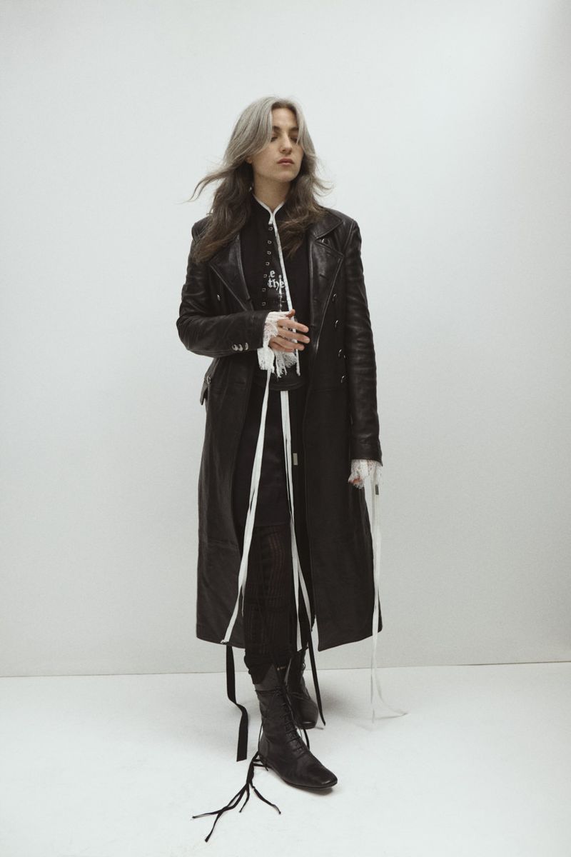 Full-length shot of a young adult woman with silver-streaked dark hair, wearing a long black leather coat, in a studio setting against a white background.