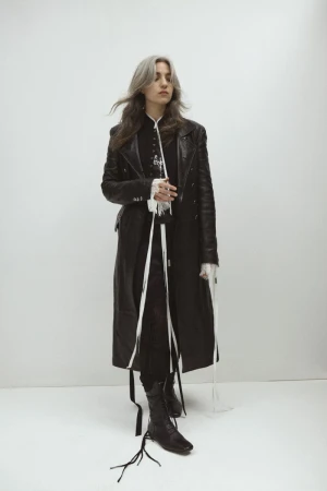 Full-length shot of a young adult woman with silver-streaked dark hair, wearing a long black leather coat, in a studio setting against a white background.