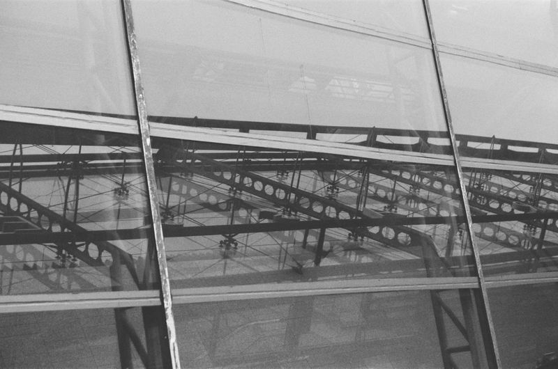 Black and white photo of industrial steel girders and supports framed by glass windows, creating an abstract architectural pattern.