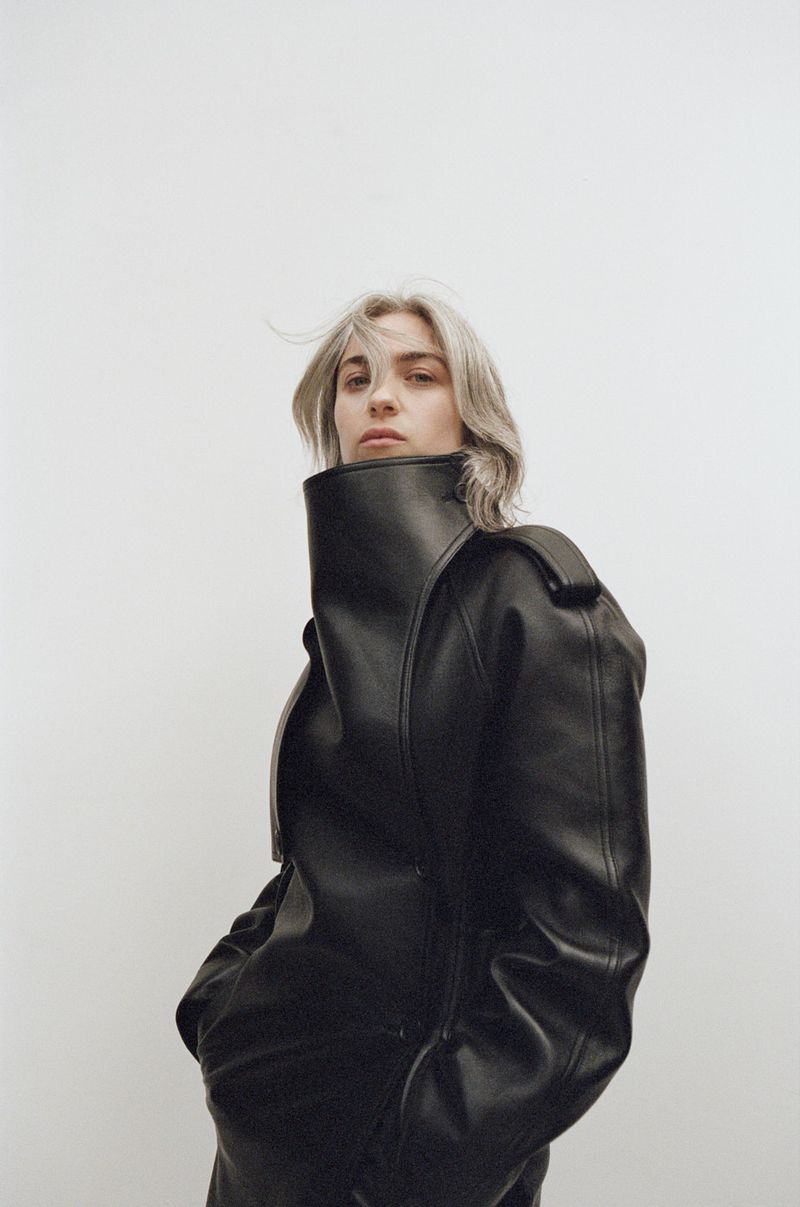 Young woman with ash blonde hair poses in an oversized black leather jacket against a neutral background in a studio-style portrait.