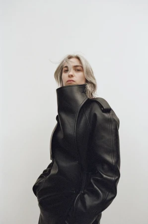 Young woman with ash blonde hair poses in an oversized black leather jacket against a neutral background in a studio-style portrait.