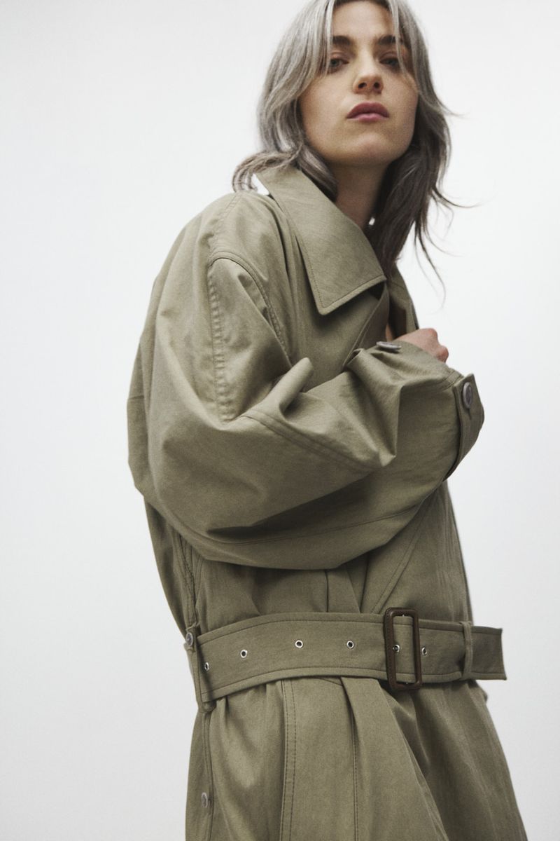 A woman with silver hair is posing, wearing a long, belted, khaki-colored trench coat in front of a plain white backdrop.