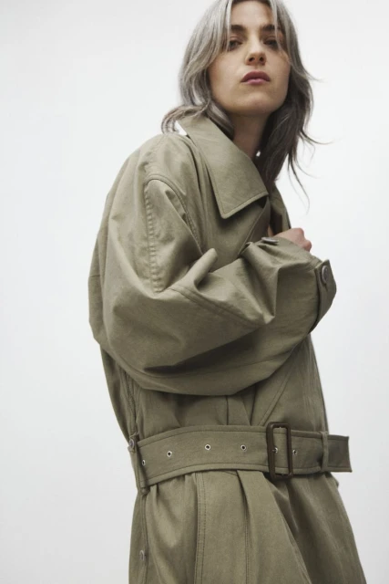 A woman with silver hair is posing, wearing a long, belted, khaki-colored trench coat in front of a plain white backdrop.