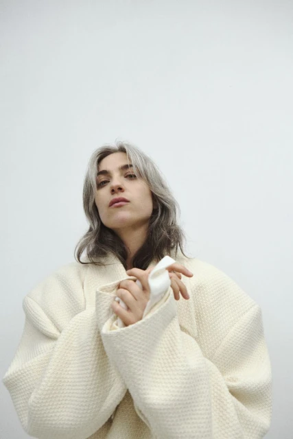 A woman with silver hair wearing a textured white coat looks directly at the camera against a neutral background in a studio setting.