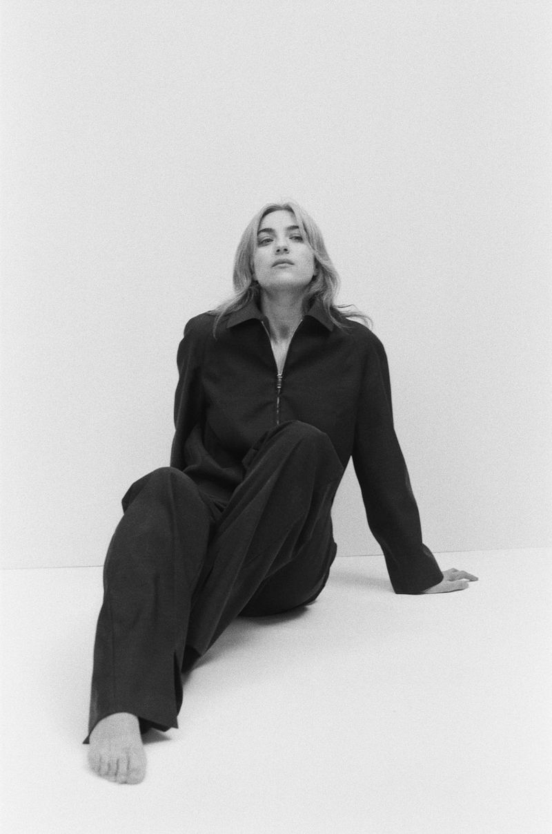 Black and white portrait of a woman in a zip-up jacket and pants sitting on a white surface, looking at the camera with a neutral expression.