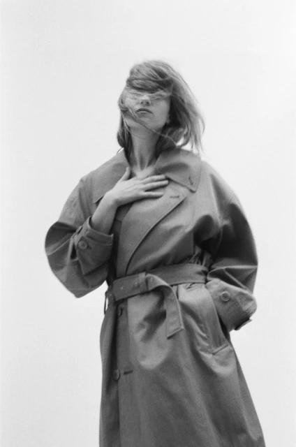Black and white fashion shot of a woman in a trench coat with hair blowing in her face, against a bright sky background.