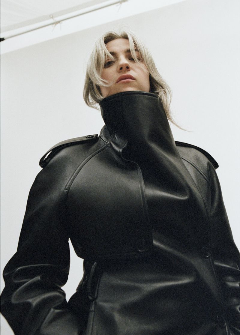 Woman in a black leather trench coat posing from a low angle with a neutral expression in a photo studio.