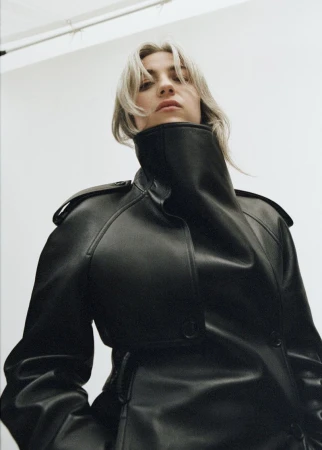 Woman in a black leather trench coat posing from a low angle with a neutral expression in a photo studio.